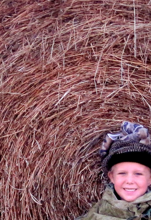 A mother holding a hunting rifle with her two young daughters crouching next to a bay of hale in a green field. All three are in camouflage.