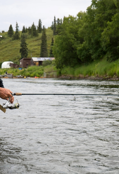 Man in fishing gear boots fishing in a river with hill of green grass and trees.