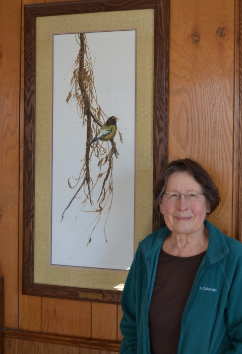 Artist Marion Otnes stands next to her framed and matted watercolor of an evening grosbeak