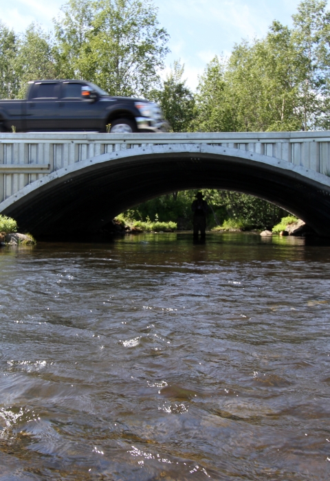 A black truck drives over a newly-constructed and much larger culvert or fish passage over a small river in Alaska.