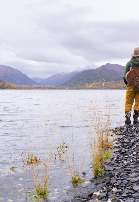 woman with a guitar stands on a lake shore looking out across the lake