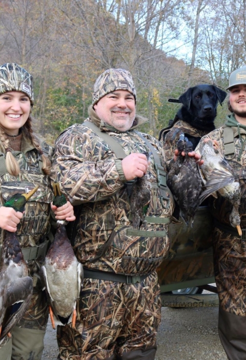 A woman and 2 men dressed in camouflage hold different species of ducks in front of their boat
