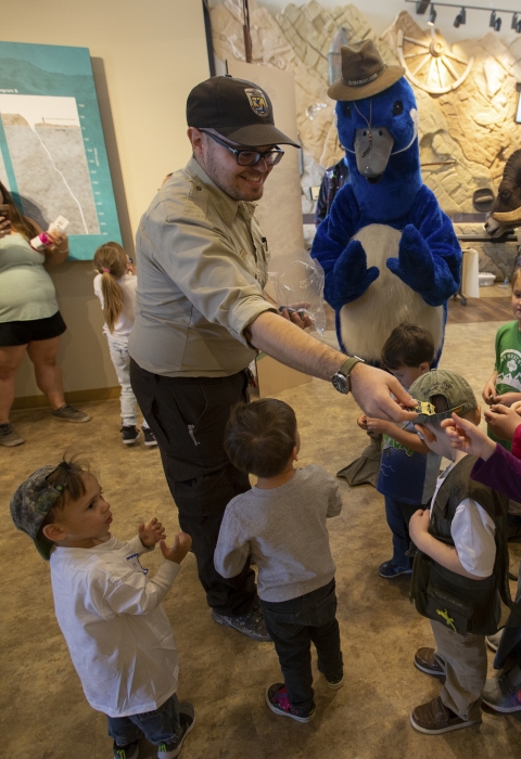 A smiling uniformed refuge ranger hands junior ranger badges out to a group of children. 