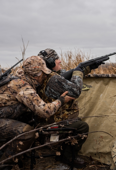 Person is camouflage hunting gear crouches behind a second person in a wheelchair in a duck hunting blind