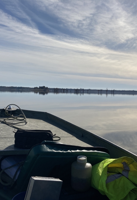 The prow of a boat looks out over a glassy lake surface reflecting cloudy sky and forests on the horizon.