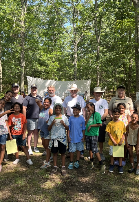 a large group of kids and adults smile in front of an archery range in the forest