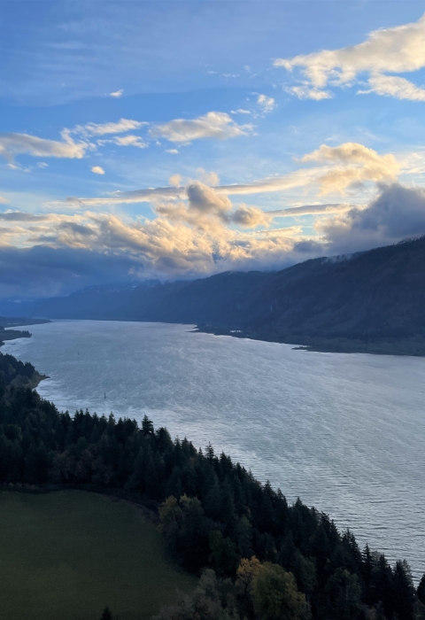 A view of a river from a high point, looking down at the river between hills and mountains with trees. The sky is blue with clouds and low, golden light.