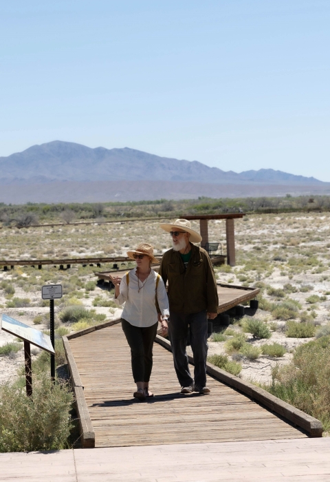 Two people walk down a raised boardwalk in a desert environment.