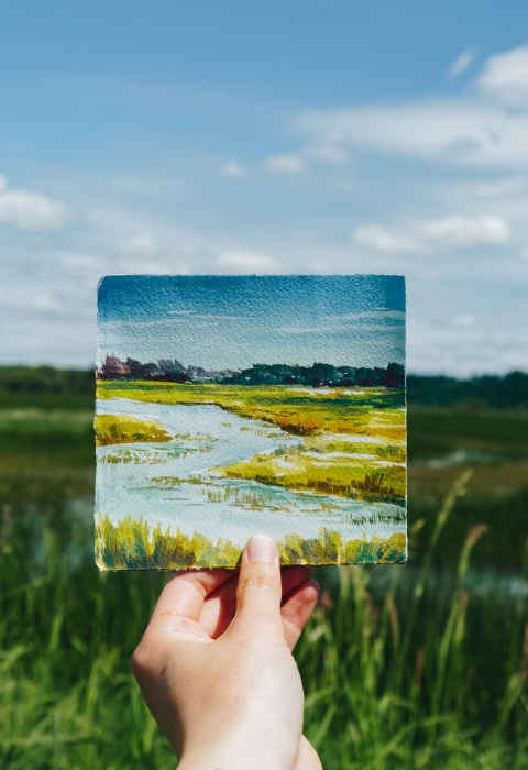 A hand holding up a tiny watercolor of the scene behind it