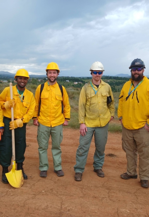 Six firefighters stand in a line for a photo. They are all in yellow nomex and fire pants. They are standing on a dry road with dry vegetation behind them.