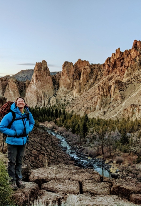 A woman wearing a blue puffy jacket and backpack stands smiling in front of a scenic view of rock cliffs and a river.