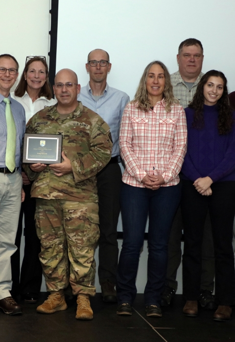 Croup of people, some in military camouflage uniforms, pose for a group photo. A man in a military camouflage uniform holds a plaque
