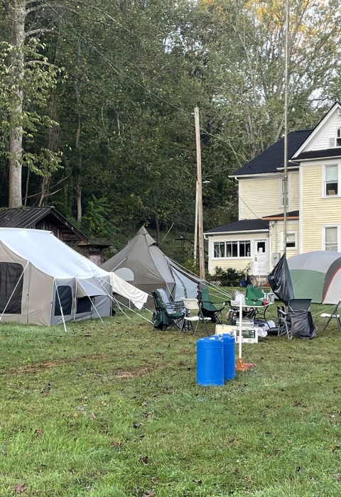 Looking across a green lawn with trees in the background is a large yellow house with a wrap around porch. In the foreground, near the house are several tents set up providing a place for storm volunteers to stay. 