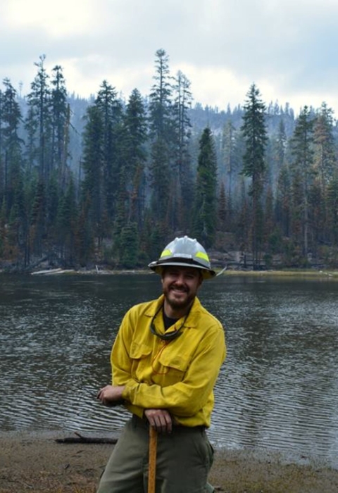 A firefighter dressed in Nomex protective clothing and hard hat, smiles and leans on their tool. Behind them is a lake and tall evergreen trees. The sky is hazy with smoke.