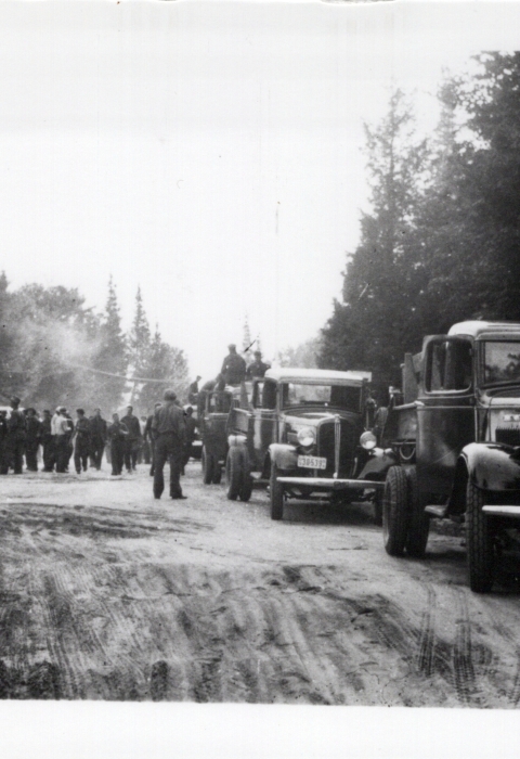 A group of men standing in a parking lot with three old fashion vehicles parked along the side of a forested road. The photo is black and white and landscape oriented. It was taken in the 1930s or 1940s.