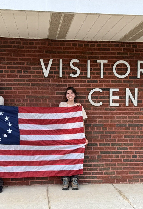 2 people hold up Betsy Ross flag in front of brick wall saying Visitor Center