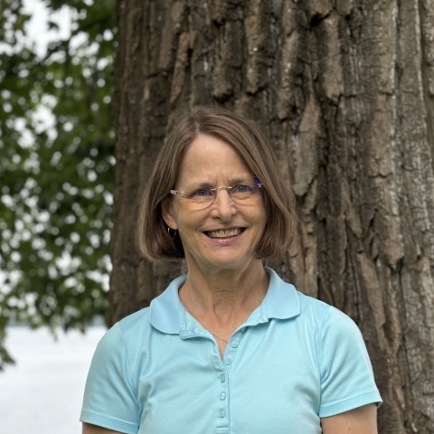 a white woman with blond hair and blue eyes, wearing glasses and a light blue polo shirt, in front of a tree, with a lake in the background
