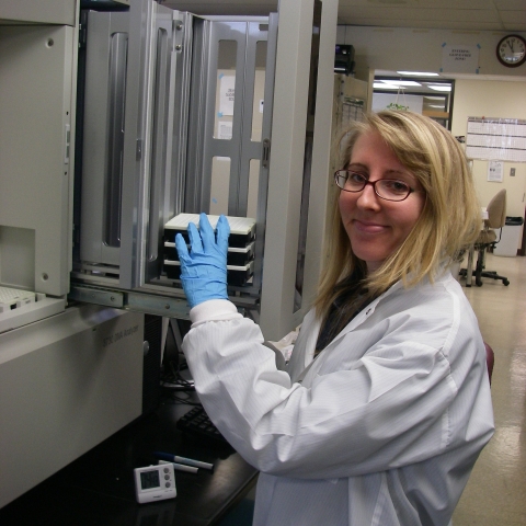 A molecular geneticist in a conservation genetics laboratory wearing a lab coat poses with some samples near some equipment. 