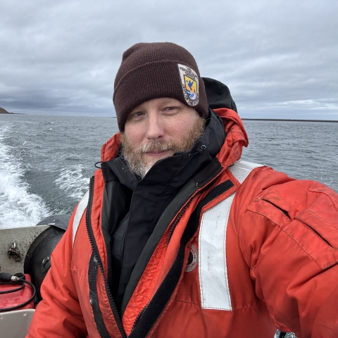 A USFWS staff member in a high visibility float coat personal floatation device and a USFWS Refuge uniform beanie takes a selfie near the stern of a boat. Open water in the background. Some land on the horizon can be seen. 