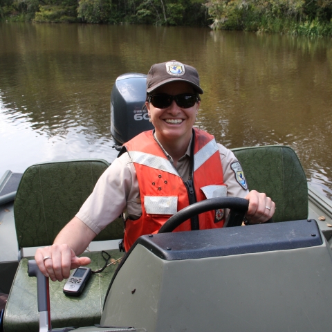 Service employee wearing an orange lifejacket driving a jon boat with an outboard motor. 