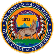 Round seal with yellow-orange background, title around the edge, and in center, a howling wolf, the outline of the reservation with the date "1872," and an eagle headdress