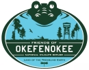 The Friends of Okefenokee National Wildlife Refuge Logo colored green and blue with an alligator, trees, dragonfly, and tower with the words "Land of the Trembling Earth" at the bottom.