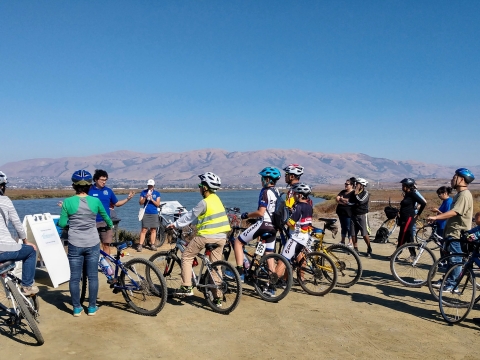Bicyclists wearing helmets gather near a refuge staff person talking about the refuge's salt marsh habitat.