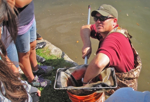 A man standing in a river puts his hand in a net, which is in a bucket, while kids stand nearby