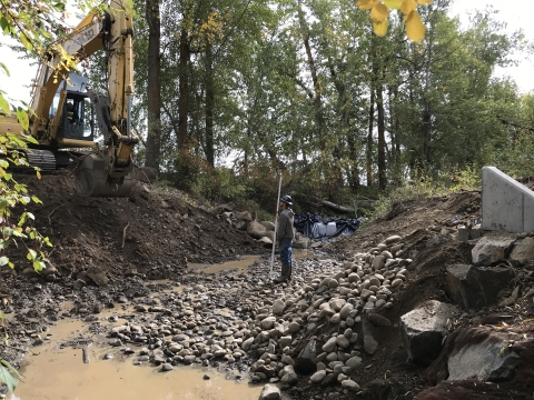 A man stands in a muddy, dewatered streambed looking up at an excavator perched on the bank.