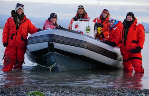 Six people in orange survival suits stand around an inflatable skiff in the water.