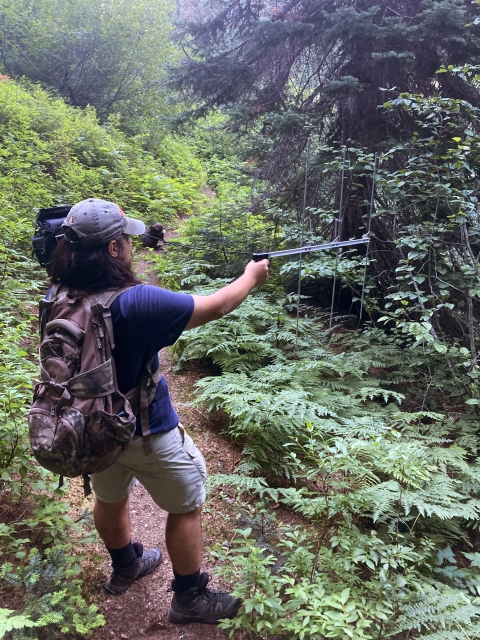 A man in t-shirt and backpack holds out an antenna while standing on a forest trail.