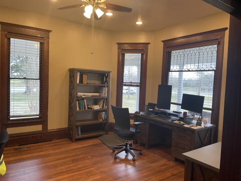 A bookshelf stands against a yellow wall between two windows with dark wooden trim. A desk, chair and monitors are placed against a wide front window. One of the porch columns is visible, behind the window. A ceiling fan is partially visible and lights are on. 