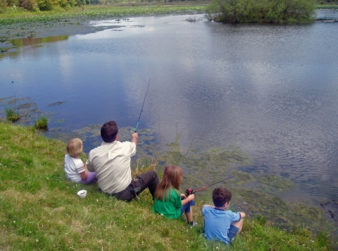 Three small children sit on the edge of a waterway with a man in a US Fish and Wildlife Service uniform. The group is fishing. 