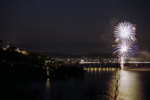 Fireworks explode over a lit up city. In the foreground a river reflects the lights of the fireworks and city, 