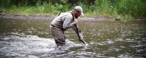 Man in light-colored clothing, hat, and fishing waders uses a stick to help him cross a knee-deep river.