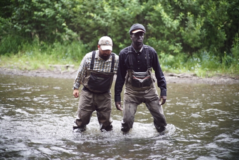 Two men in fishing waders shuffling their feet across the bottom of the knee-deep river to show how to safely cross a river.