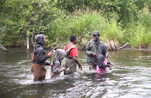A group of youth are practicing safely wading across a knee to waist-deep river while walking in fishing waders.