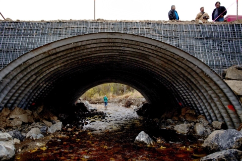 A fish-friendly culvert arch is being constructed out of cement and metal, showing someone walking at the far side of the culvert in the creek, as well as three men working on the road side of the culvert.