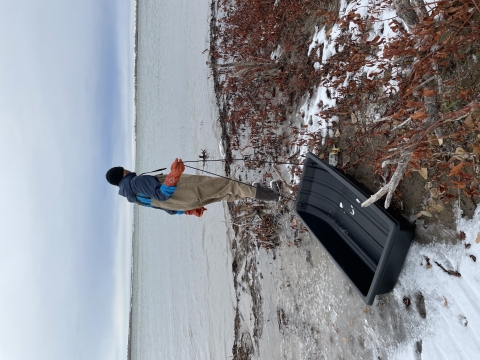 A person dressed in waders, gloves, and other protective equipment pulls a sled along the lakeshore.