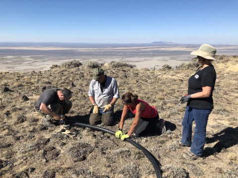 People work together on to replace a water pipe on a ridge, with a valley distant in the background. 