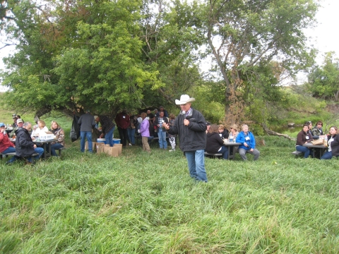 A man wearing a cowboy hat stands on a grassy field, speaking to a crowd of people seated around small tables nested in a copse of trees