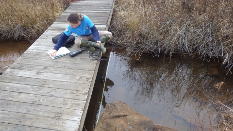 AmeriCorps intern sitting on a wooden bridge writes down water quality data she measured with a YSI.