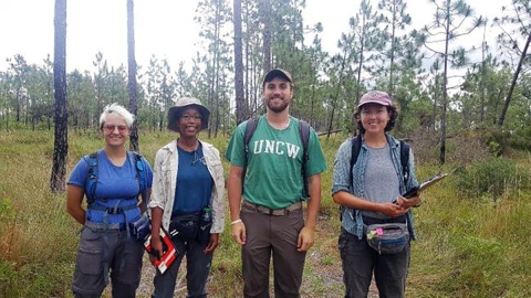Four people standing smiling for the camera with pines and grass in the background.