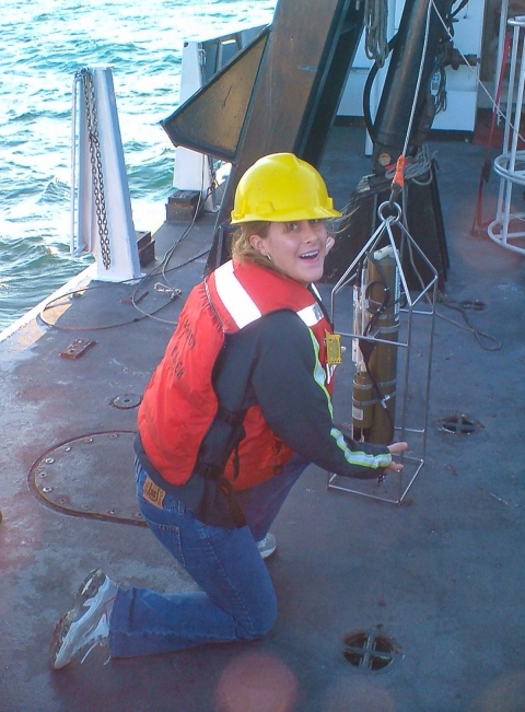 A woman wearing an orange life vest and yellow hard hat smiling and crouching the deck of a ship at sea.