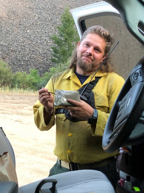A man in fire clothes eats an MRE meal along side a truck
