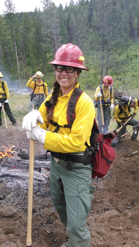 A woman stands smiling wearing a red helmet, bright yellow shirt, green pants, and a red backpack. Behind her are a few people dressed similarly working with a small smoldering fire.