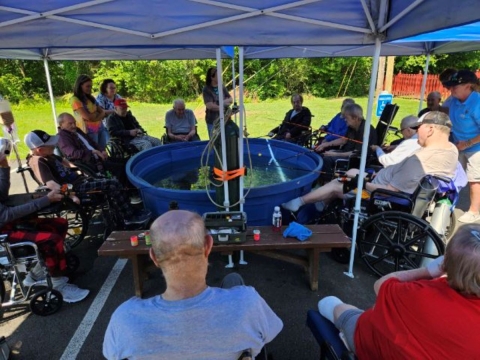 Elderly people gathered around a large blue tank with fishing poles