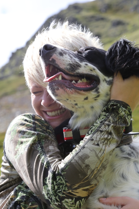 Christine Cunningham hugs her dog colt while smiling. Colt the Dog has his mouth open and tongue slightly sticking out. Out of focus hill in the background. 