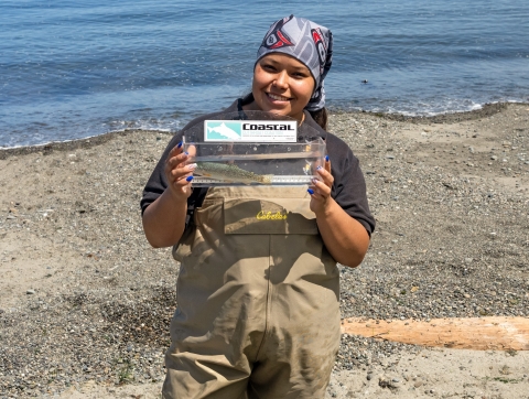Service Tribal intern standing along a shoreline holding a fish in a small tank.