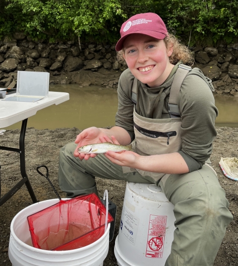 Service intern, Kathleen Gerard, holds a coastal cutthroat trout while seated. Fish sampling buckets and equipment are in view around her and water and vegetation are in the background.